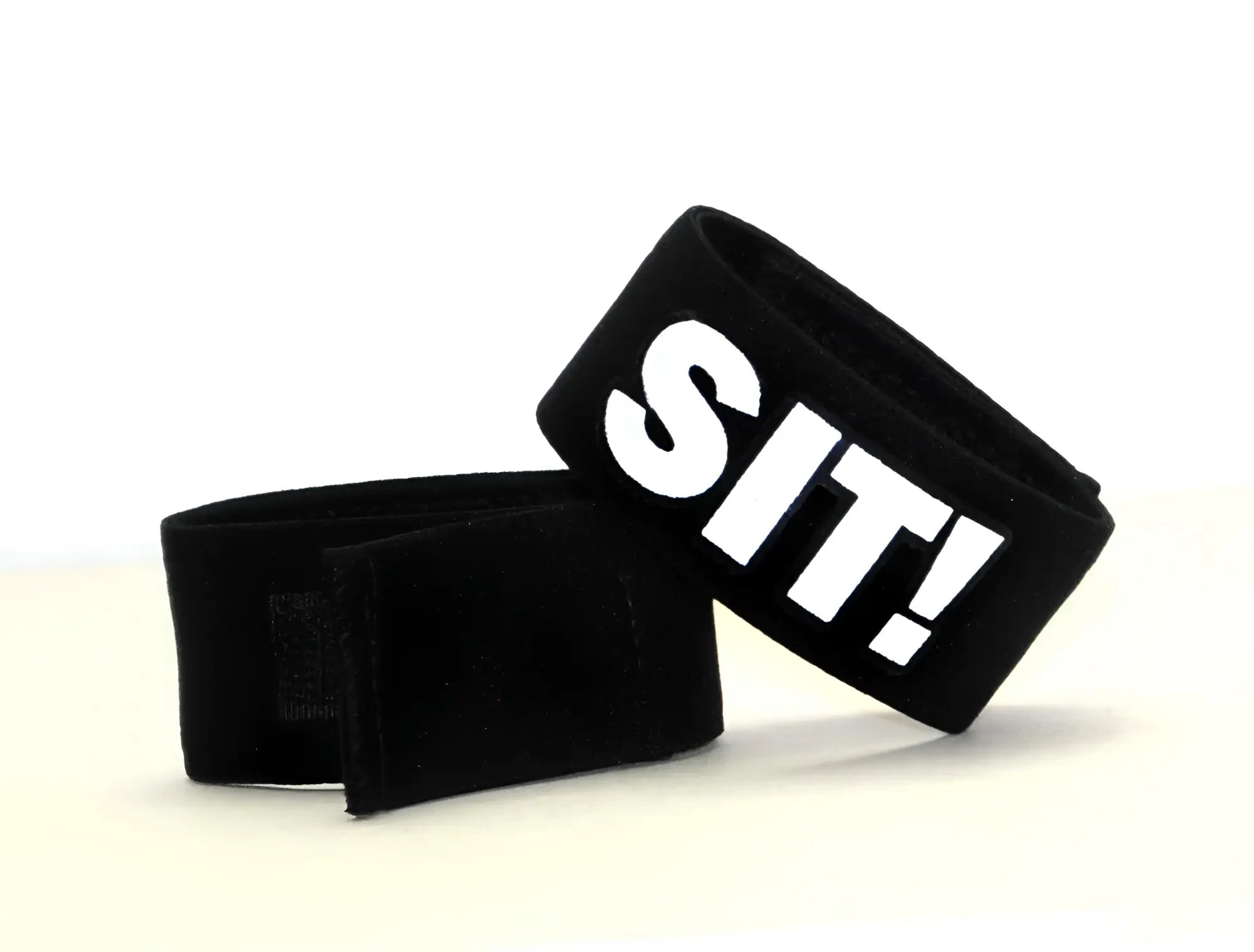 Two black fabric cock-ring bands with velcro closures, one featuring the white text "SIT!" on a white background.