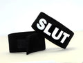 Black fabric cock ring with white "SLUT" lettering and Velcro fastener.