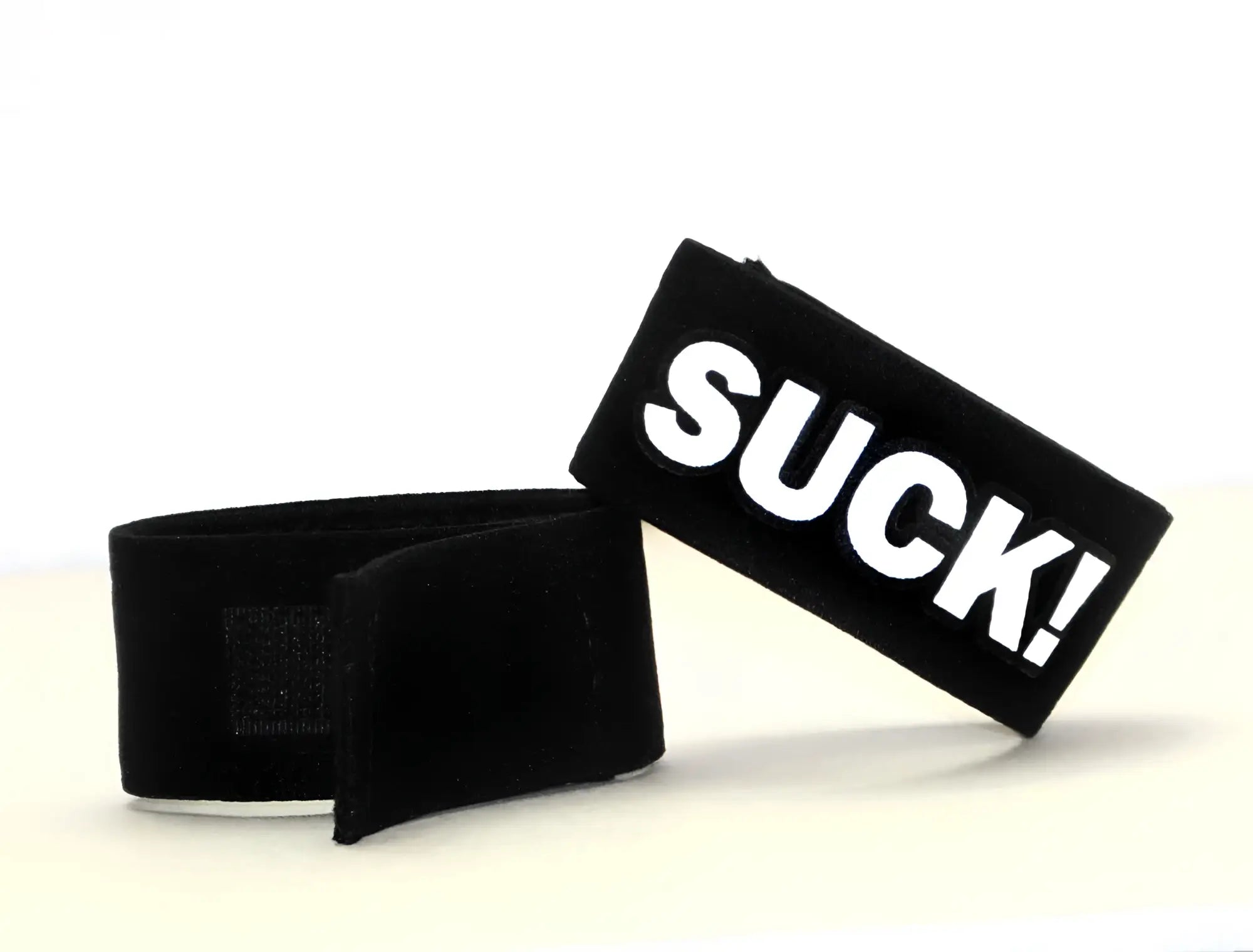 Black fabric cock ring with a Velcro closure and a white patch featuring the bold black text "SUCK!"