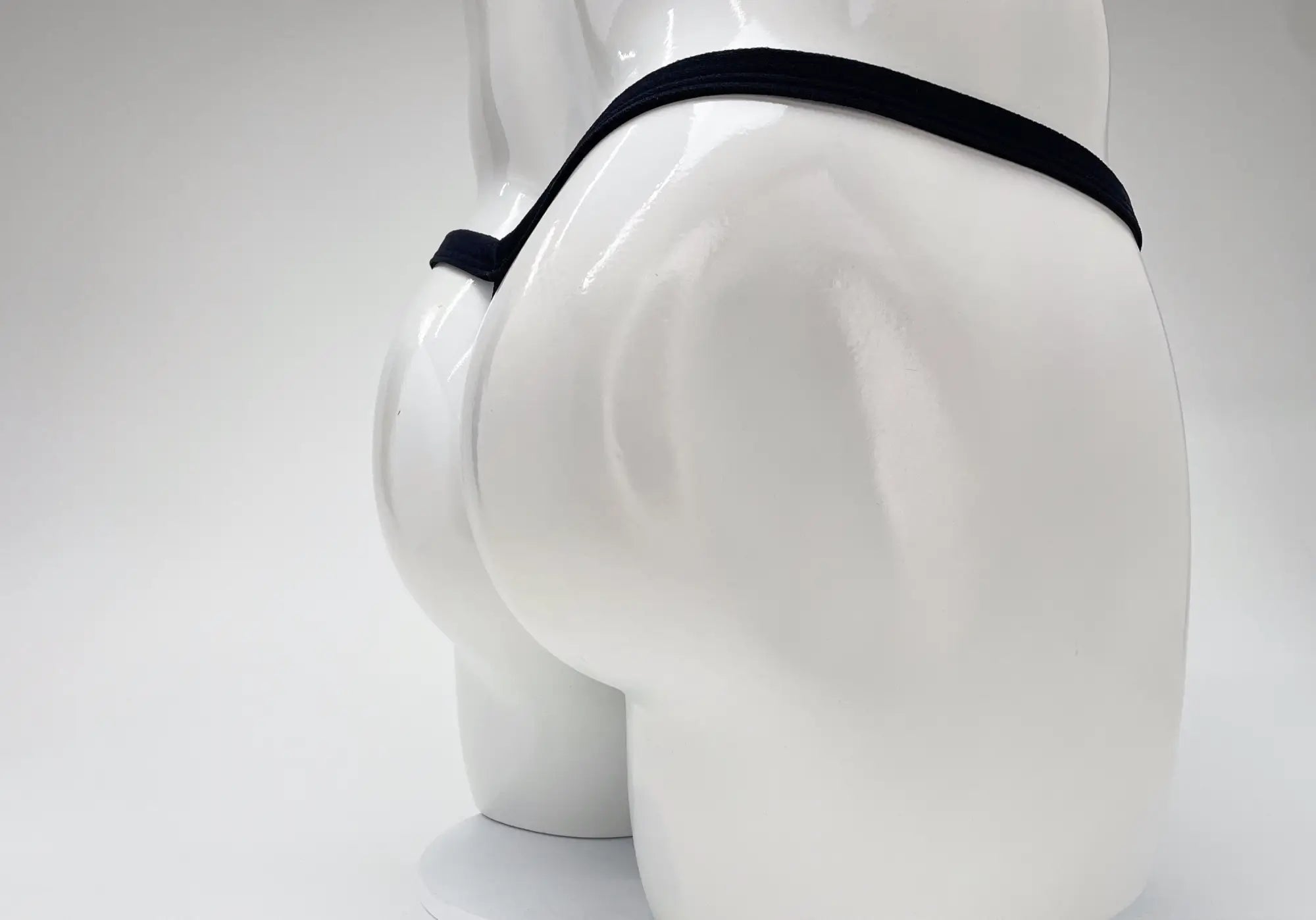 A back view of a white mannequin wearing a thin, black micro thong with a minimalist strap design.