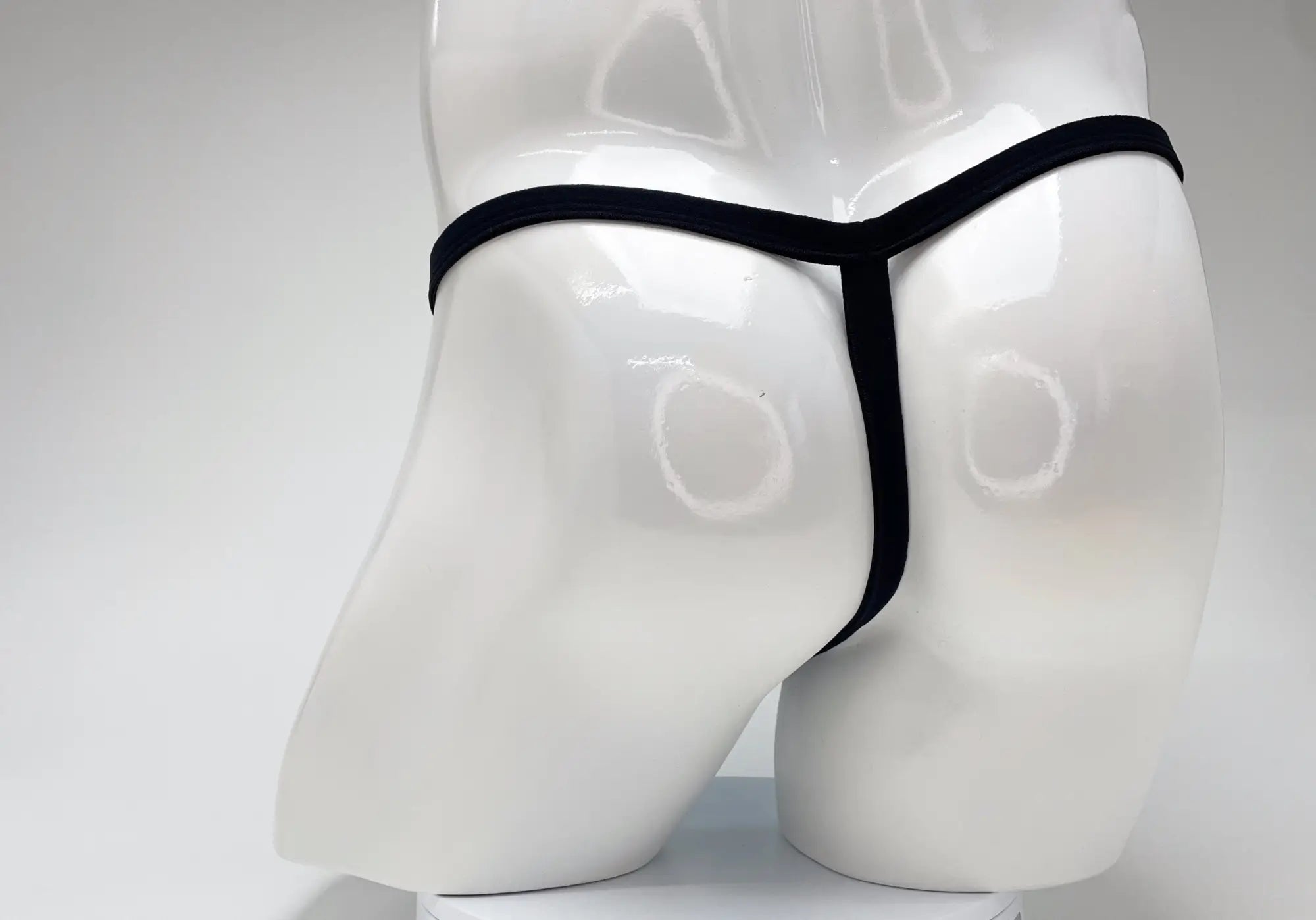 Back view of a black thin-strap thong on a glossy white male mannequin torso.