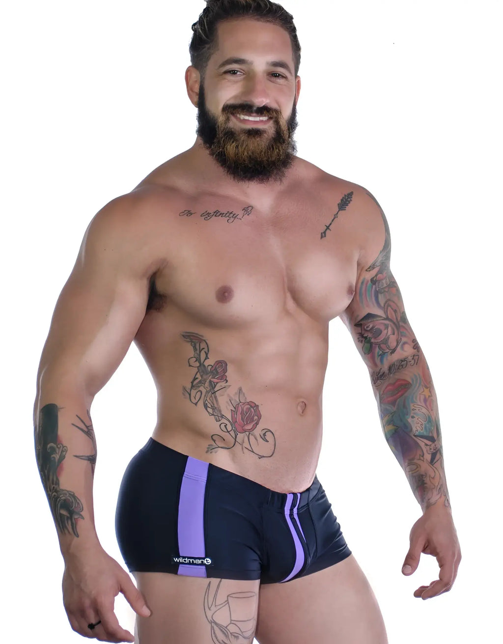 A bearded man wears black square-cut swim trunks with vertical purple stripes and a Wildman! logo on the side.