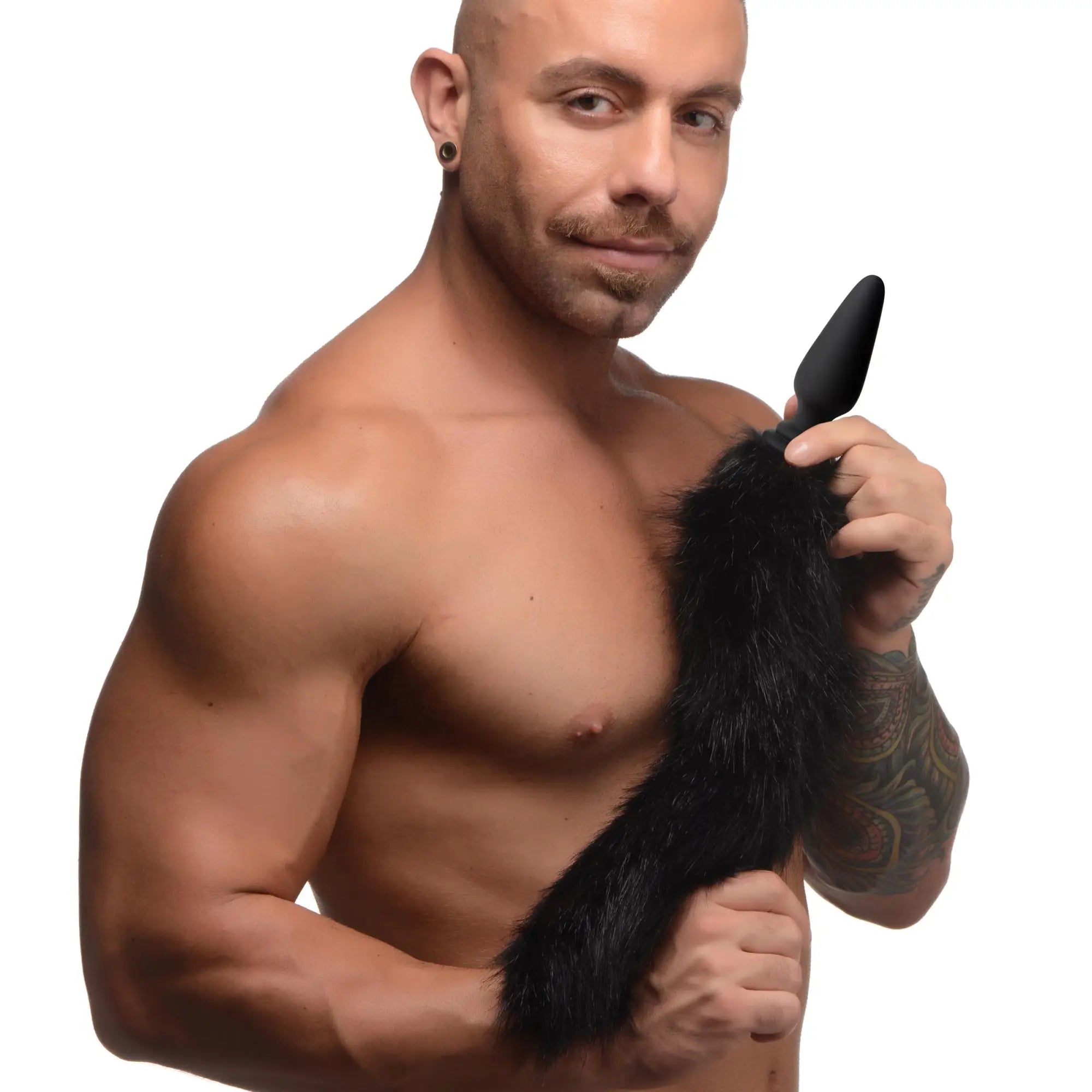 A shirtless man holding a black XR Brands interchangeable silicone anal plug with a fuzzy black faux fur tail attachment.