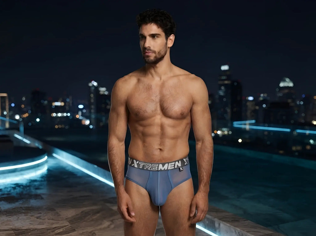 A man wearing Xtremen blue sheer briefs with a branded waistband against a blurred city skyline at night.