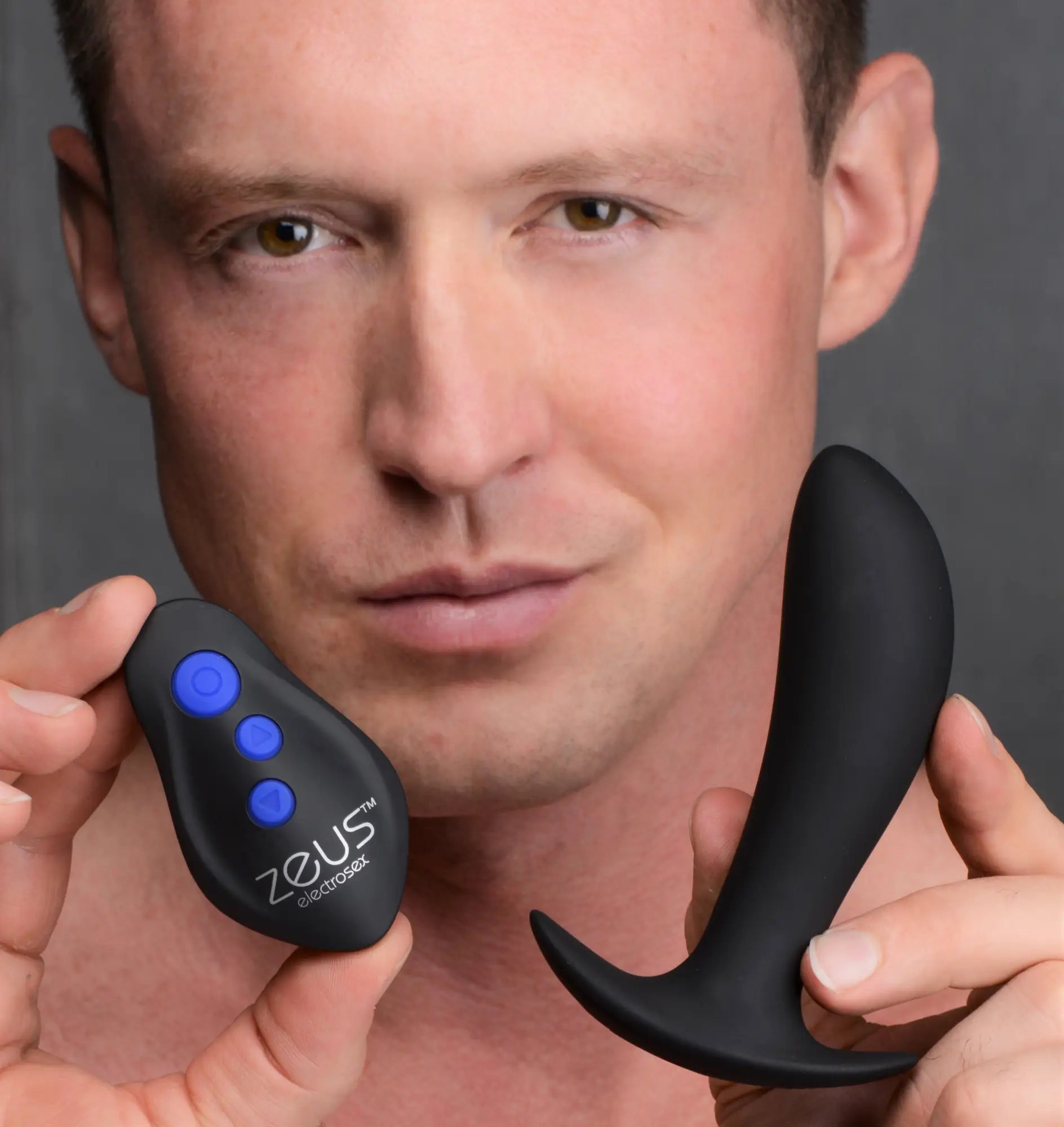 A man holds a black Zeus Electrosex 64X Pro-Shocker vibrating prostate plug and its matching remote with blue buttons.