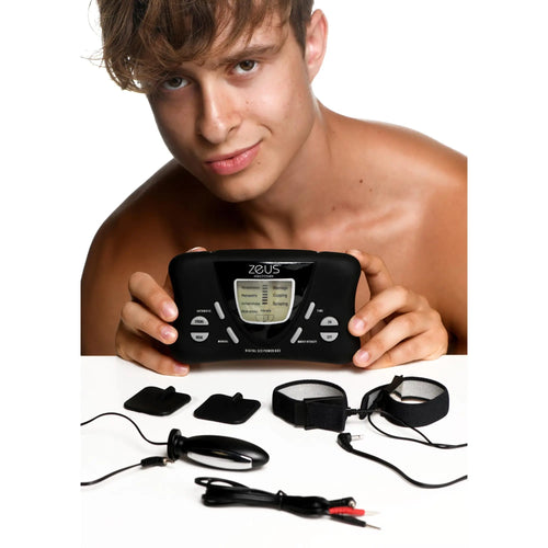 Man holding a black Zeus Deluxe E-Stim device with digital screen, electrodes, wrist straps, and cables on white table.